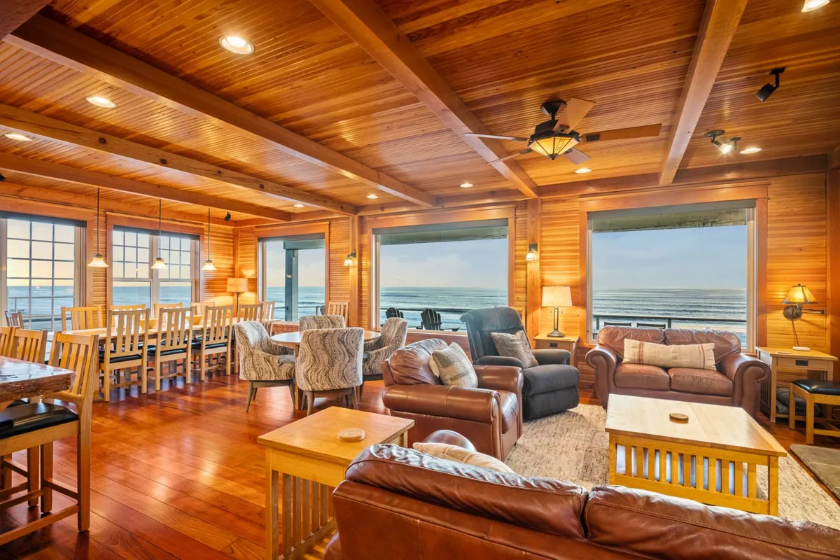The image shows a spacious, inviting interior with rich wood paneling and a large dining table, all oriented toward expansive windows that frame a breathtaking ocean view. A cozy seating area is arranged for relaxation, and the soft natural light adds to the warm, welcoming atmosphere.
