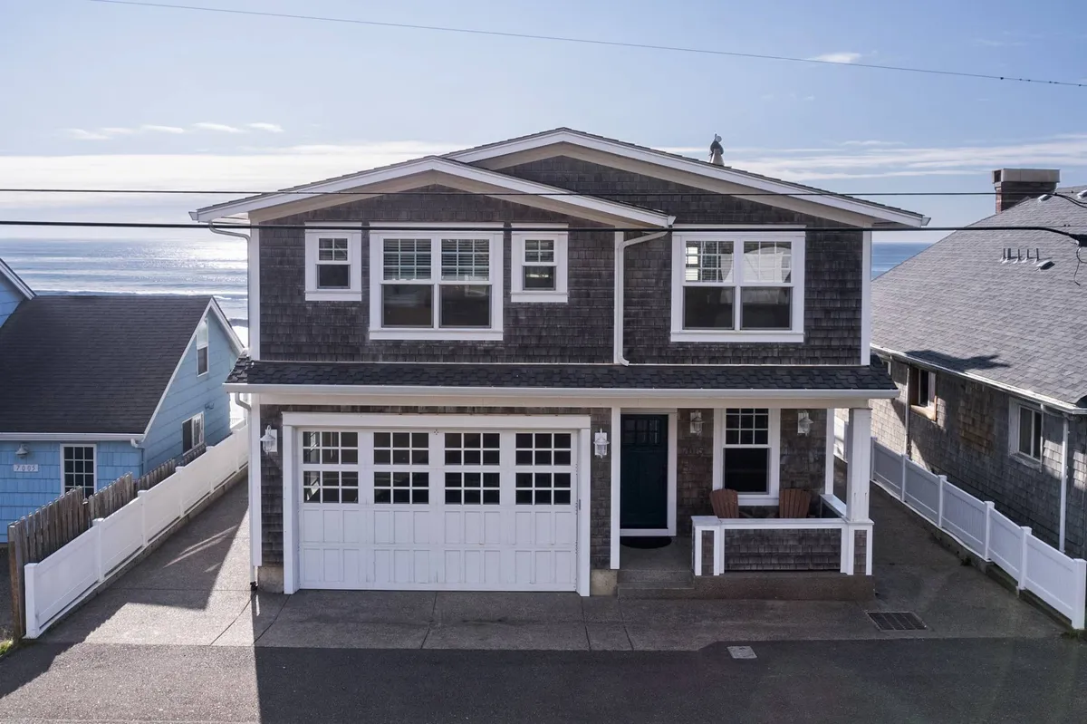 This image showcases a welcoming two-story house with a garage door and front porch, set against a backdrop of the ocean. The exterior looks inviting and serene, suggesting both comfort and direct access to beautiful coastal scenery. The setting is ideal for gatherings, relaxing on the porch, and enjoying ocean views, which connects perfectly to the spirit of local exploration.