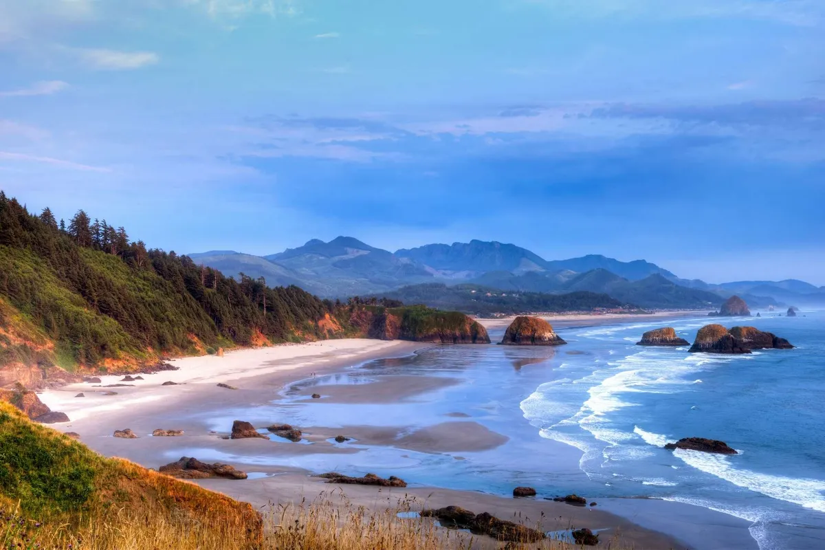 This scenic view captures the rugged coastline, sandy beach, distant mountains, and crashing waves, offering a glimpse of the outdoor activities available nearby such as hiking, beachcombing, and tide pool exploration. The setting is vast and inviting, perfect for adventurous guests.