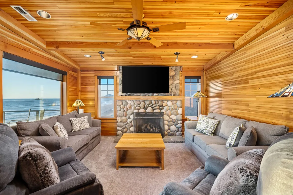 A spacious, warmly lit living room featuring a fireplace, large windows framing the ocean view, comfortable sofas, and wooden ceiling. The space feels luxurious yet welcoming, inviting group gatherings with a backdrop of natural beauty.