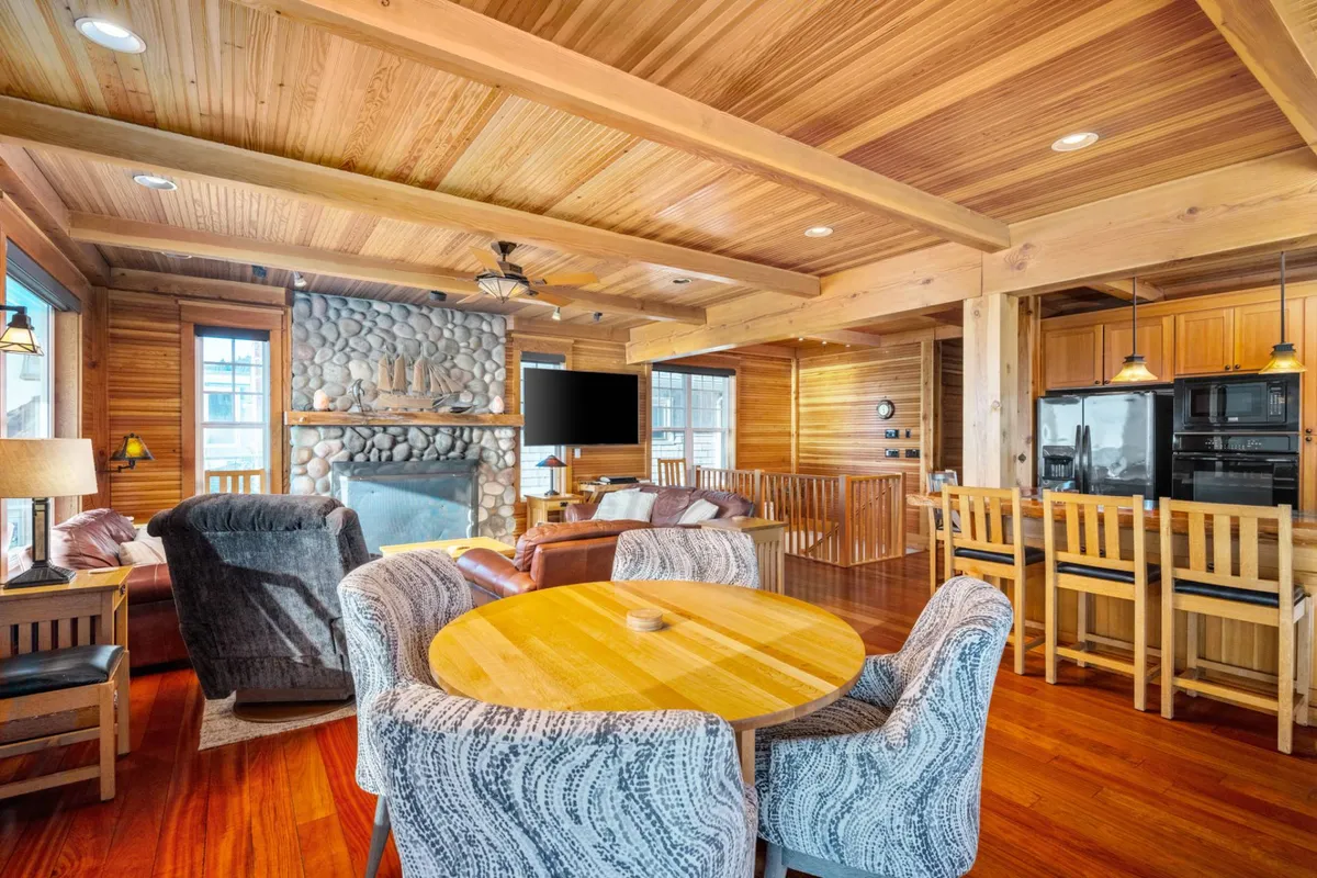 The image features a spacious open-concept area that seamlessly integrates a stone fireplace, a wooden dining table, a comfortable seating area, and a kitchen bar. Large windows flood the space with natural light, and Craftsman-style woodwork is prominently displayed throughout the interior. The harmonious blend of textures—wood, stone, and soft furnishings—creates a welcoming and communal atmosphere.