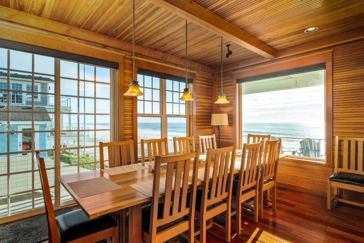 A long wooden dining table, elegantly set beneath hanging pendant lights, sits in front of large windows that provide breathtaking ocean views. The room is bathed in sunlight, creating an inviting atmosphere perfect for large group gatherings or celebratory feasts.
