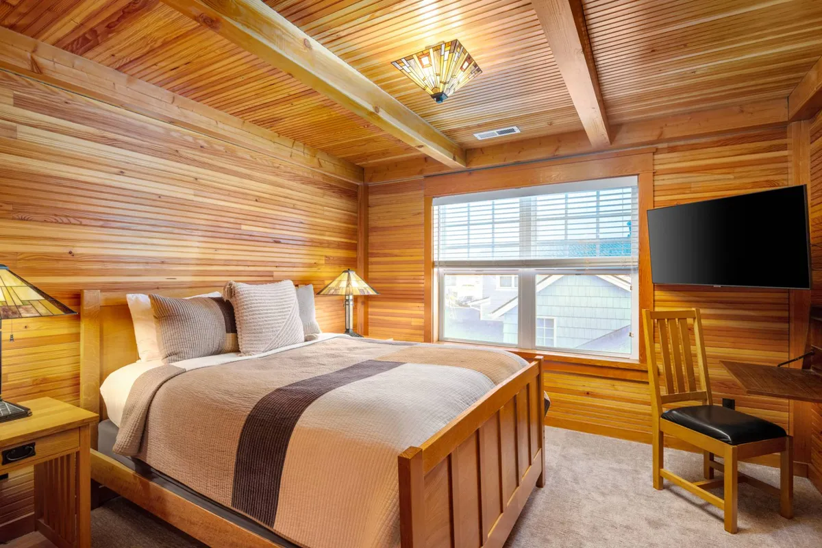 A serene bedroom with a comfortable bed, lamps, window letting in natural light, a desk for working, and a flat-screen TV. The décor blends cozy comfort with practical work amenities, and the window suggests a calming view, perhaps of the ocean.