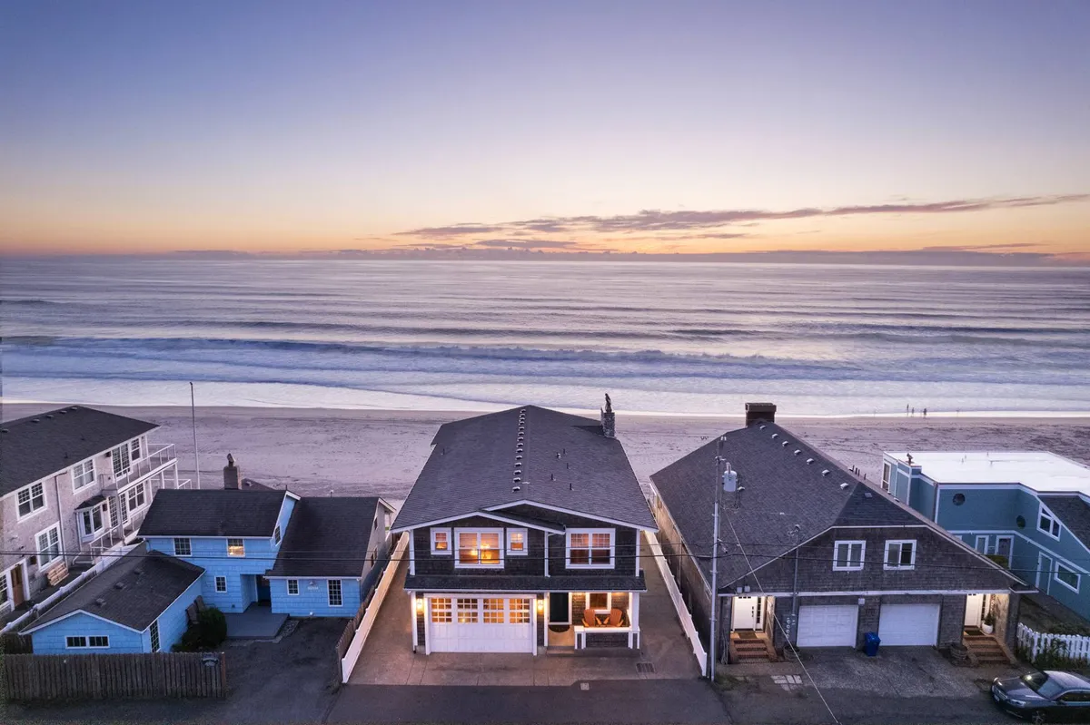 A picturesque beachside view with Sandy Shores prominently in the foreground, waves rolling in, and a dramatic sunset sky. The home’s location and the expansive beach highlight opportunities for outdoor gatherings and quiet reflection alike.