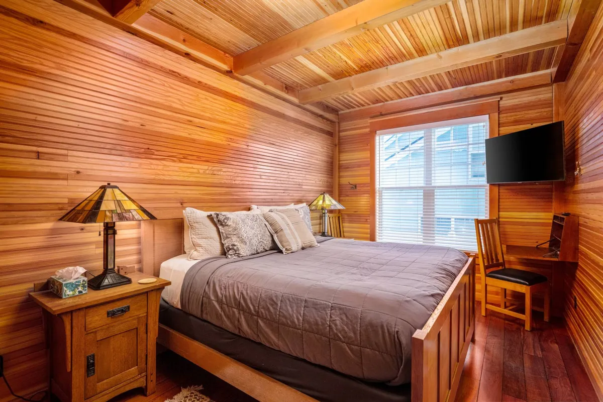 A tranquil bedroom featuring a comfortable bed with plush bedding, a bedside lamp, a window allowing in natural sunlight, a wall-mounted TV, and wooden furniture that adds warmth to the space. The overall ambiance is peaceful and homely, ideal for restful nights.