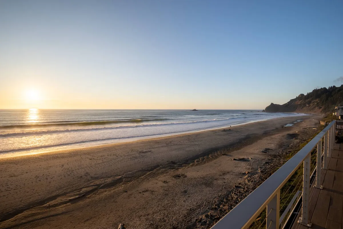 A tranquil shot of the sunset casting warm colors over the ocean, with gentle waves lapping at the shore and distant cliffs framing the horizon. The empty beach invites peaceful evening walks, beachcombing, or gathering for a bonfire as dusk settles.