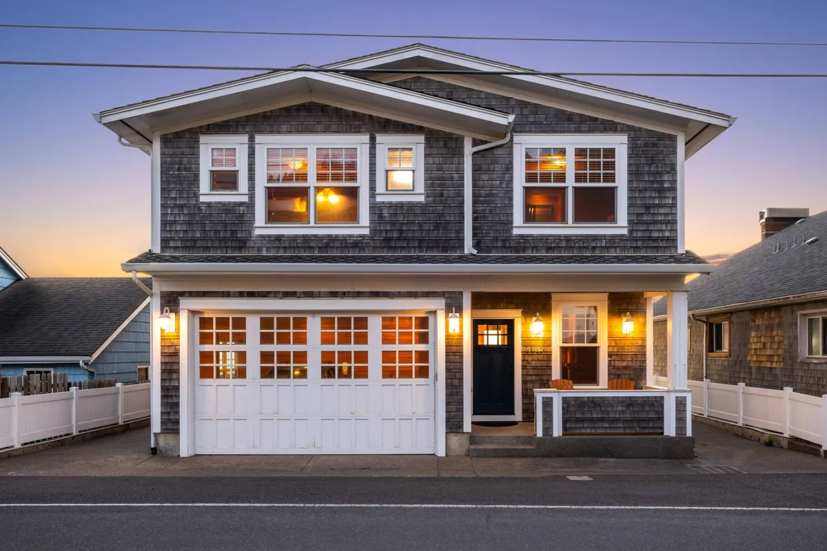 A welcoming exterior view of Sandy Shores featuring a shingled facade, brightly lit windows, a white garage door, and a charming front porch. The scene is set for gatherings, with warm lights hinting at activity inside and a spacious entrance inviting guests to connect both indoors and out.