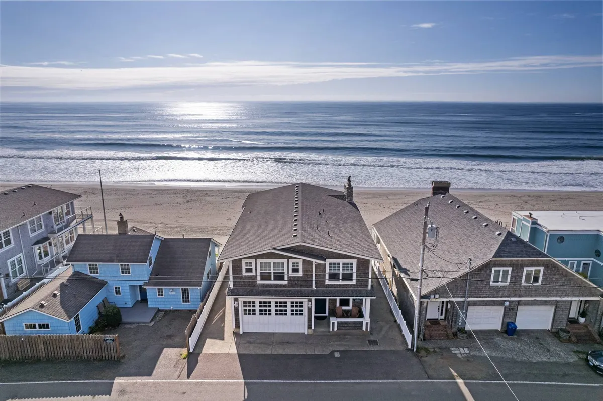 A wide view of sandy beachfront homes directly facing the ocean, with gentle waves rolling in and footprints in the sand. Families and children can be seen building sandcastles and combing the beach for shells, evoking a sense of carefree beach adventure.