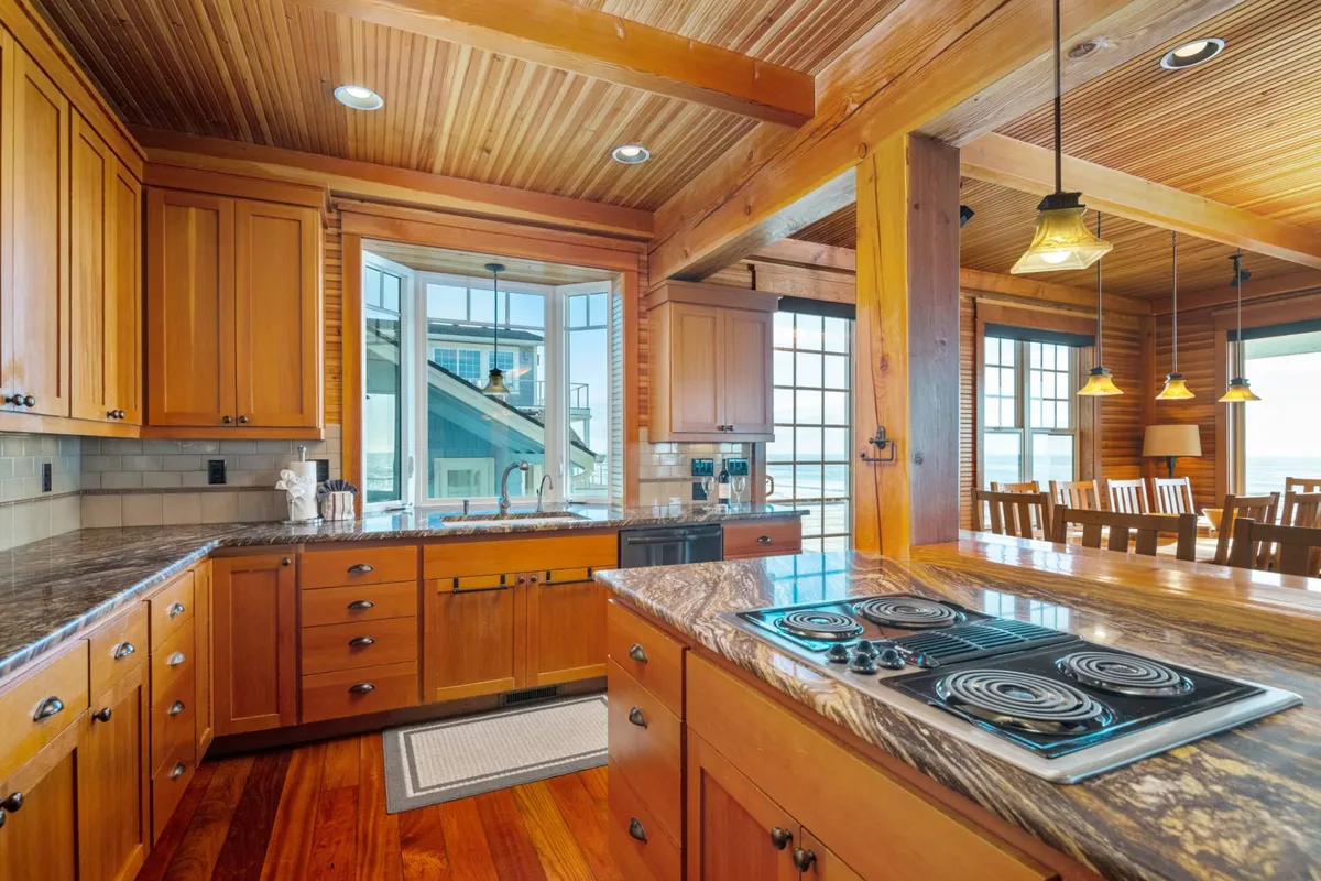 The kitchen showcases beautiful wooden cabinetry, a marble countertop, modern pendant lights, and expansive windows. The adjacent dining area is visible, and the entire space is filled with natural light, suggesting a seamless transition between cooking and entertaining.