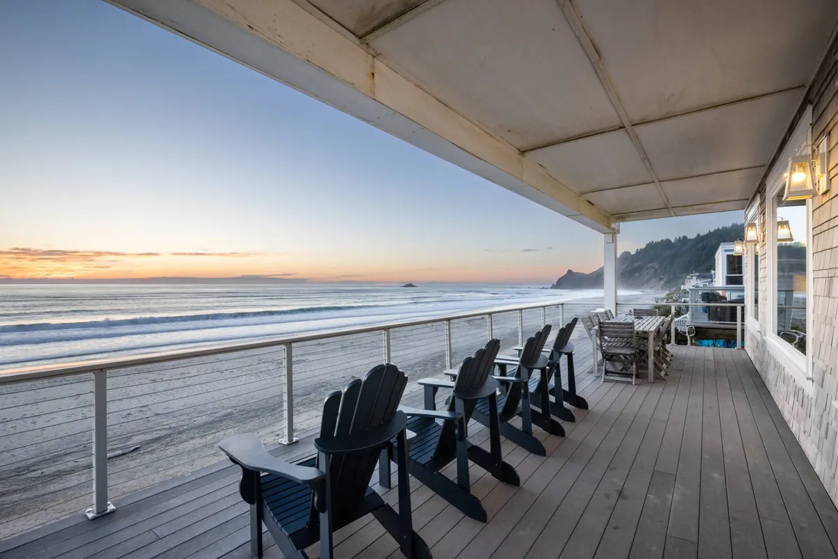 A wooden deck furnished with Adirondack chairs offers a prime vantage point for sunset watching over the ocean, with warm hues in the sky and gentle waves visible below. The tranquil setting suggests cozy evenings, group gatherings, or moments of quiet reflection against a spectacular coastal backdrop.