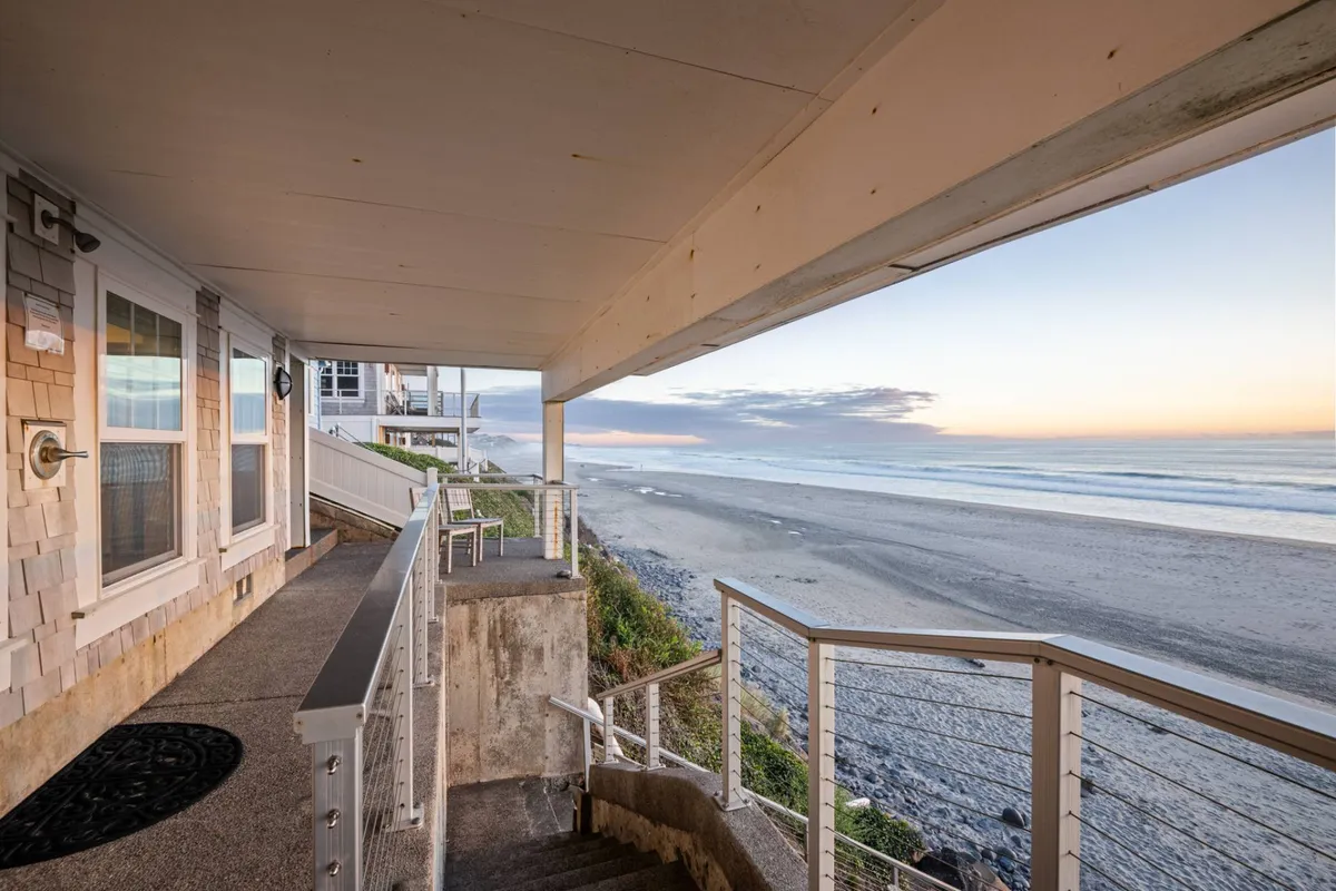 An exterior perspective showing a private staircase leading directly down to the sandy beach, with an expansive ocean view framed by soft clouds and the sun dipping toward the horizon. The scene hints at easy transitions from home to shoreline and the promise of tranquil sunsets.